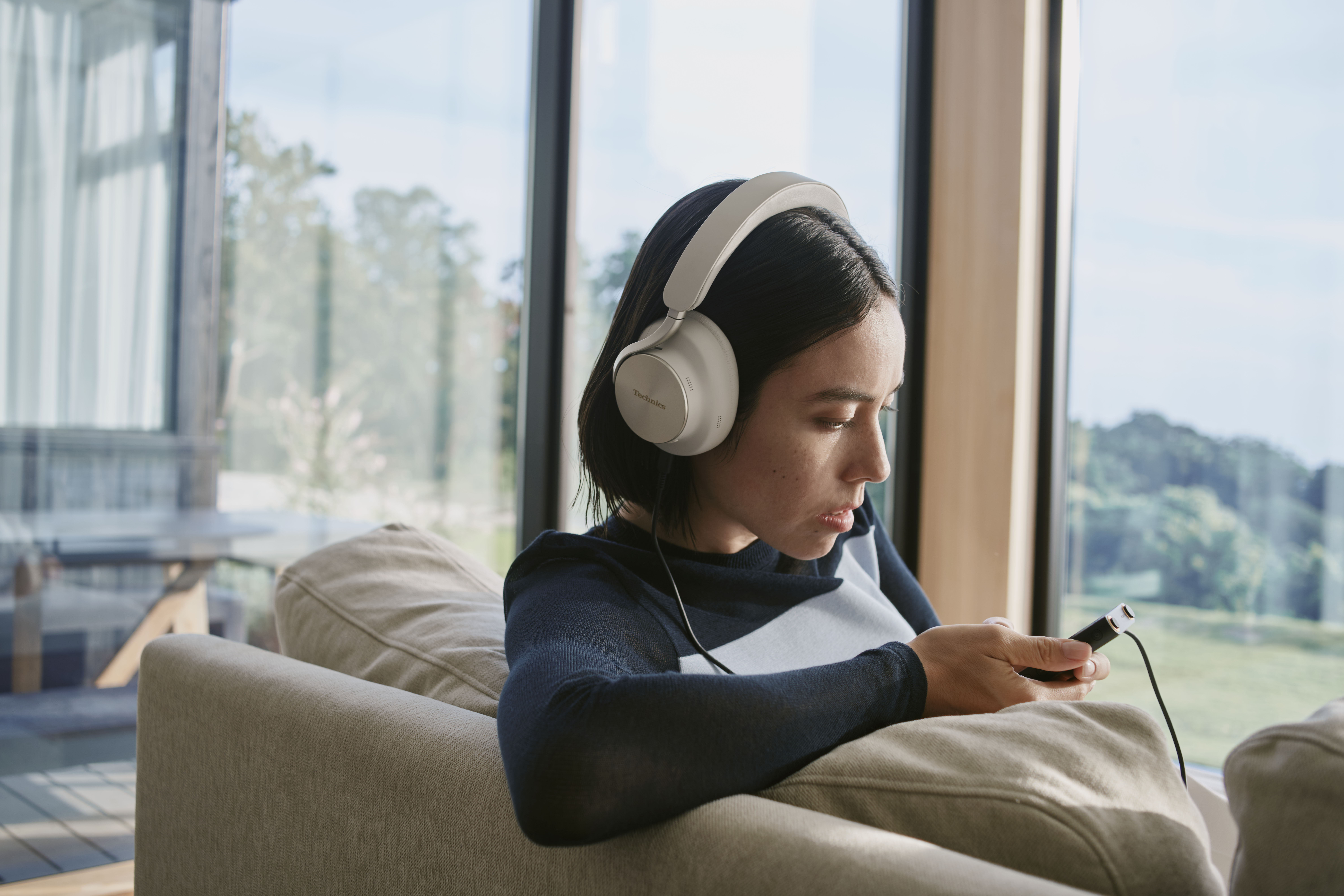 Technics EAH-A1000E-S Technics EAH-A1000 bezprzewodowe słuchawki z adaptacyjną redukcją szumów ANC (parowanie wielopunktowe Bluetooth, LDAC, do 45H pracy z ANC), srebrne 43 Kobieta słuchająca muzyki z odtwarzacza DAP za pomocą przewodu, w słuchawkach nausznych marki Technics w kolorze greige.