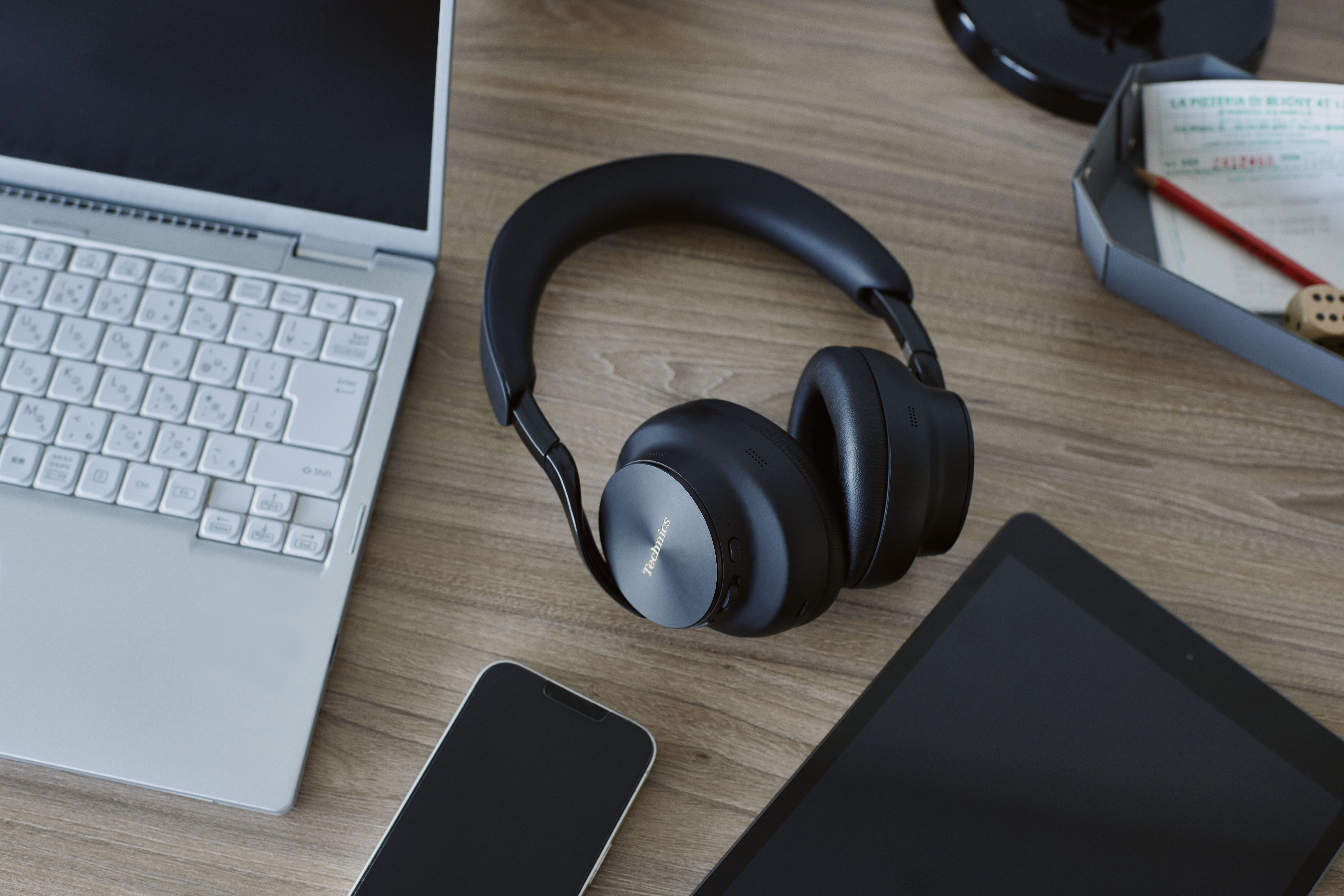 Technics EAH-A1000E-S Technics EAH-A1000 bezprzewodowe słuchawki z adaptacyjną redukcją szumów ANC (parowanie wielopunktowe Bluetooth, LDAC, do 45H pracy z ANC), srebrne 44 Czarne słuchawki nauszne marki Technics leżące na biurku obok laptopa.